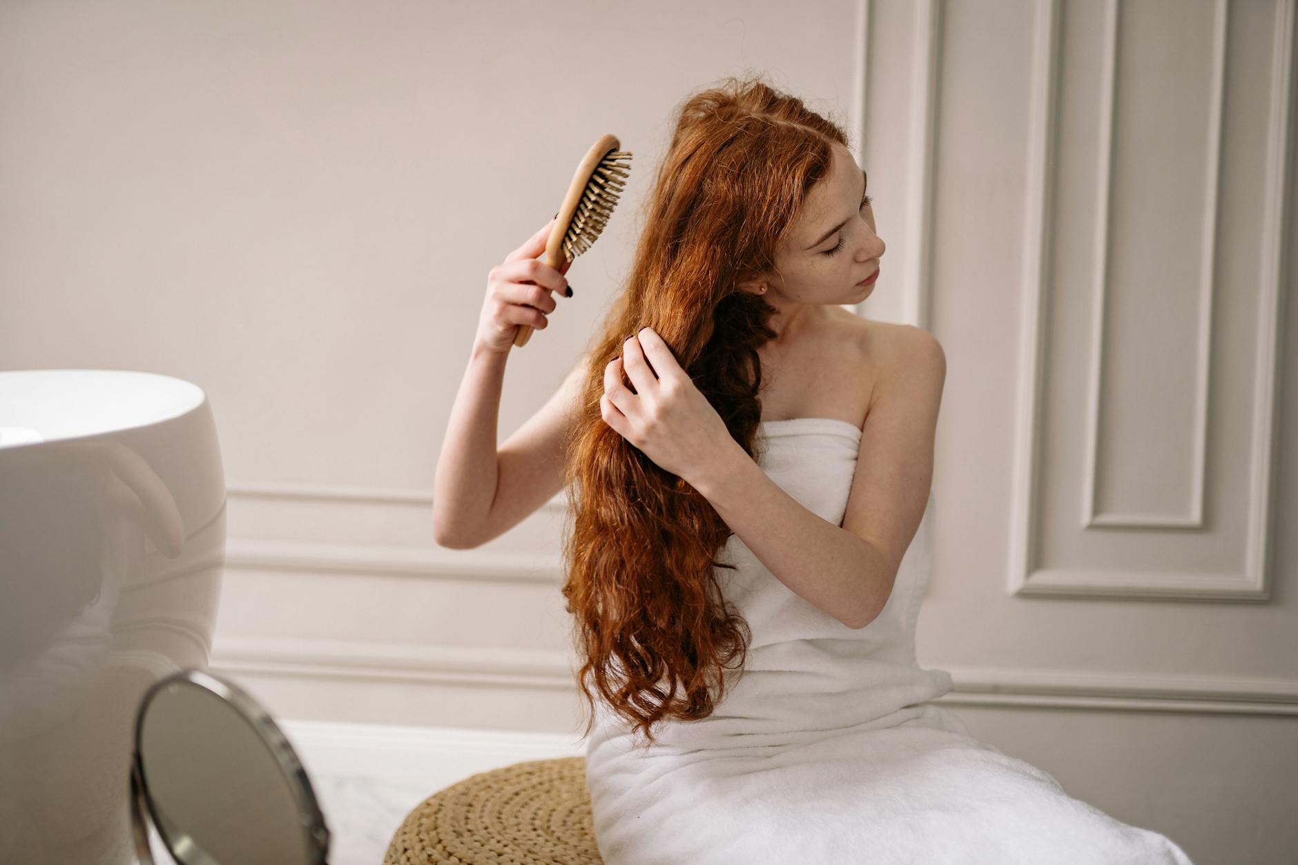 Woman with long red hair wrapped in a towel combing her hair in an elegant bathroom setting.