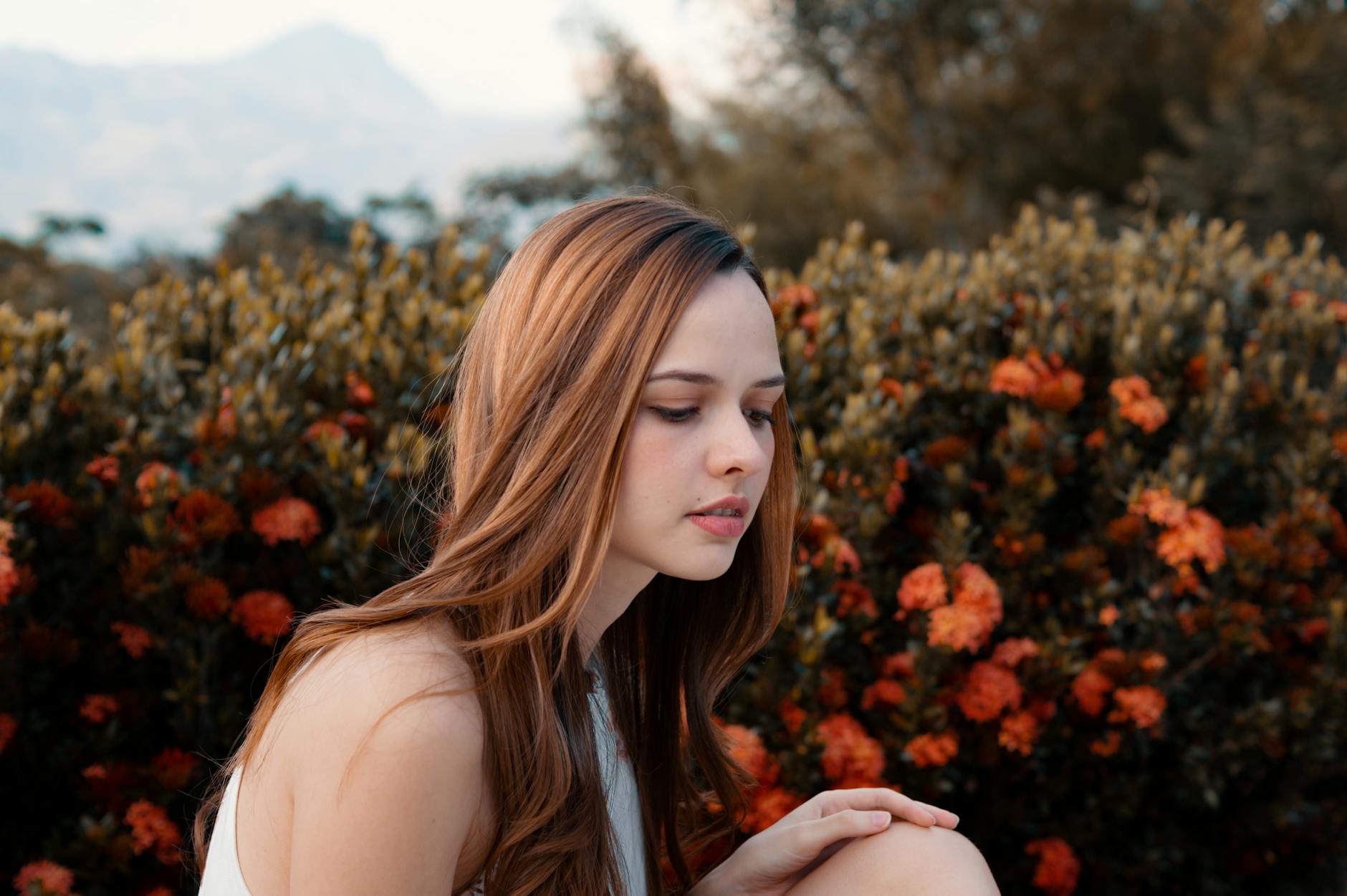 A young woman with long brown hair poses gracefully in front of vibrant orange flowers outdoors.