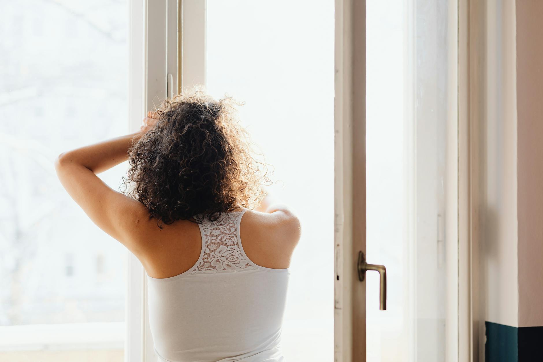 A woman enjoying a peaceful morning, standing by a window, embracing the warm sunlight.