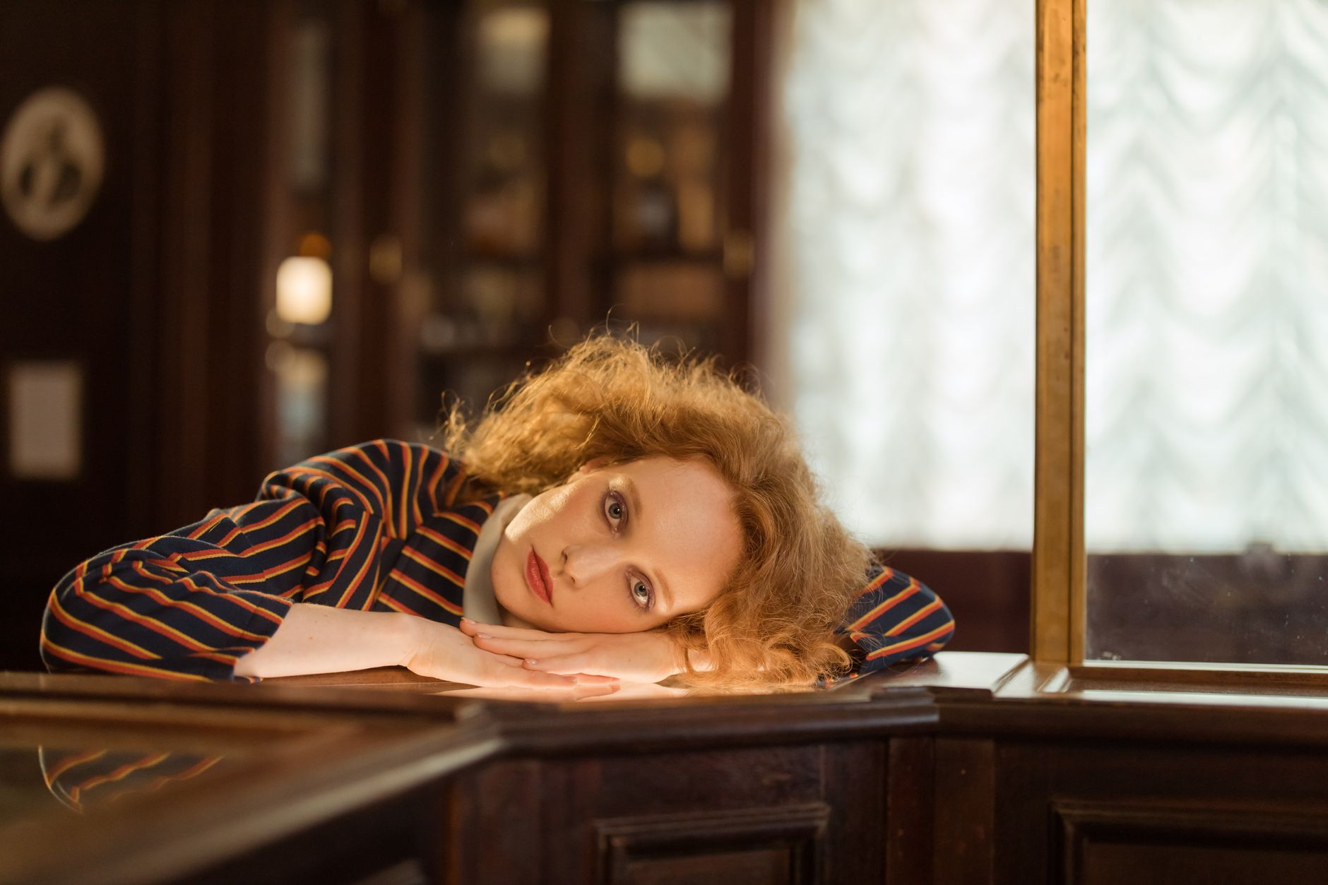 A woman with curly hair leans on a table inside a warm, ambient room.